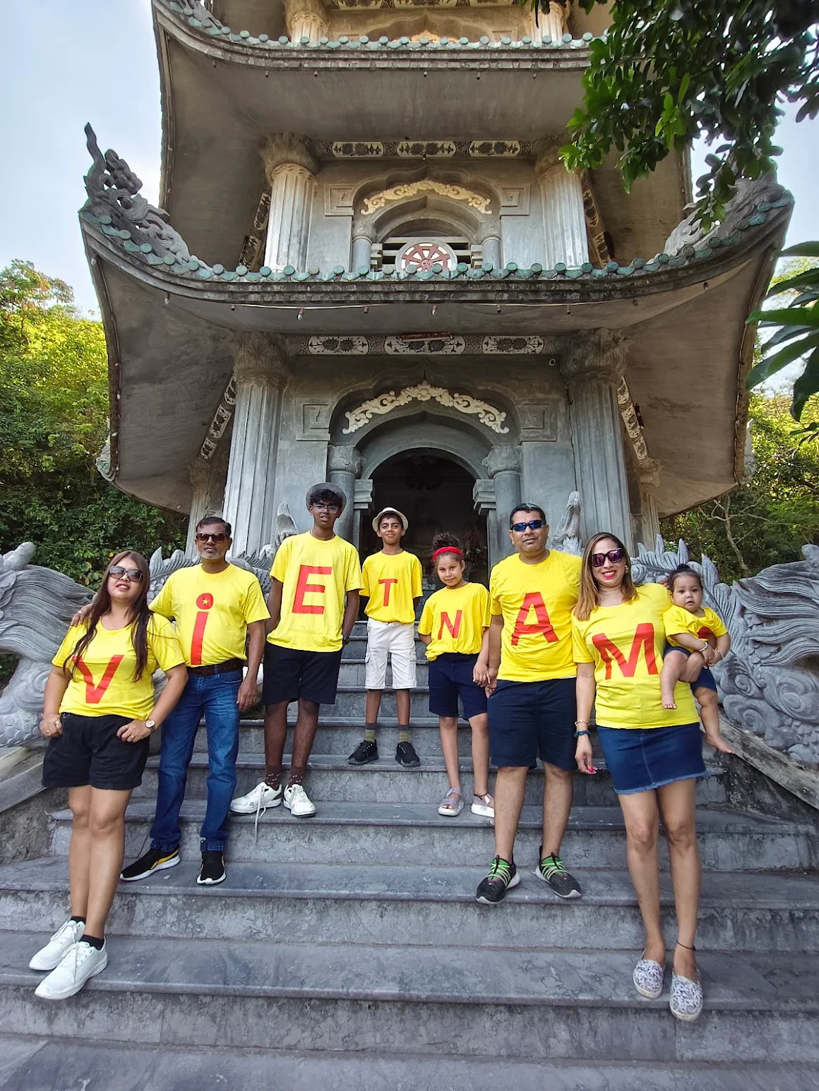 A happy family group wearing yellow shirts that spell “VIETNAM,” posing in front of a historic pagoda during their tour organized by the best travel agency in Delhi, Fab Holidays.